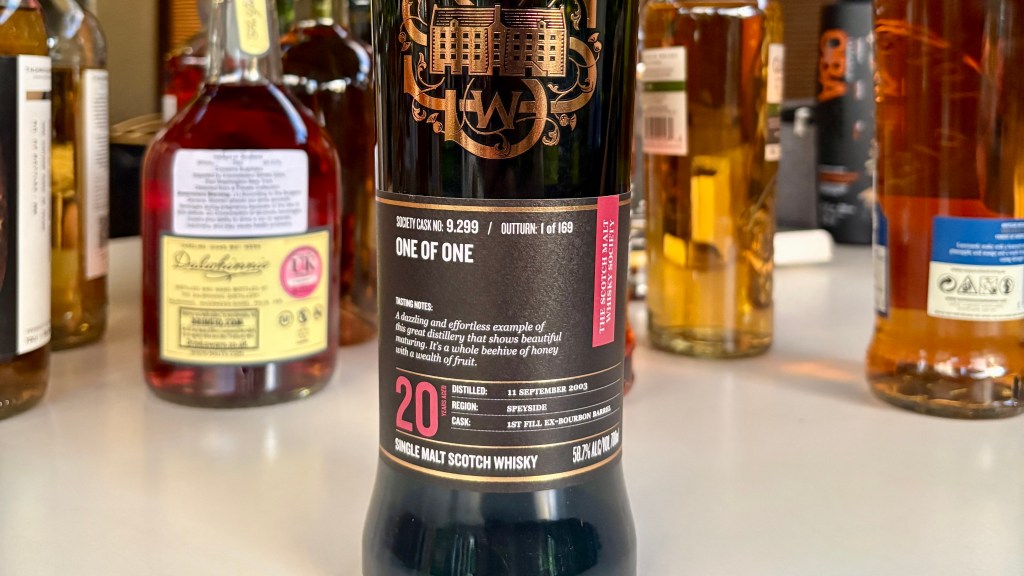 Close-up of a bottle of single malt Scotch whisky, featuring detailed label information including distillery name, cask number 9.299, and tasting notes. Background shows various other whisky bottles.